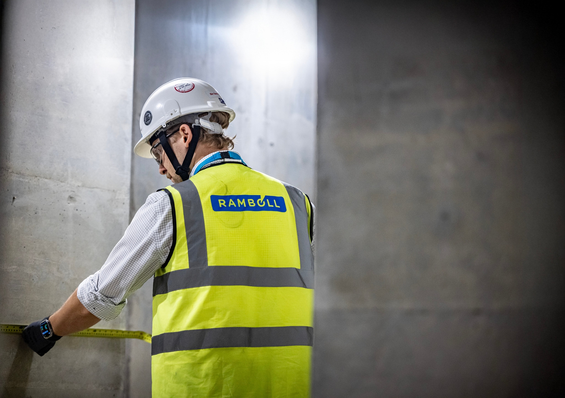 A person wearing a Ramboll-branded yellow safety vest and white hard hat measures a concrete wall in a construction setting.